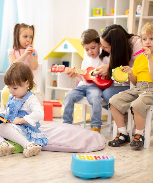 Abbildung Frau zeigt einem Kind das spielen auf einer Ukulele, weitere Kinder spielen oder lesen