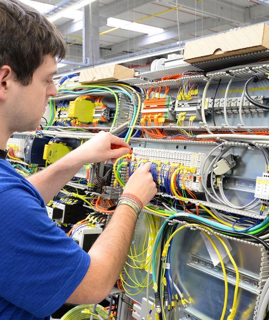Abbildung junger Mann steht an einer elektronischen Anlage und prüft Leitungen