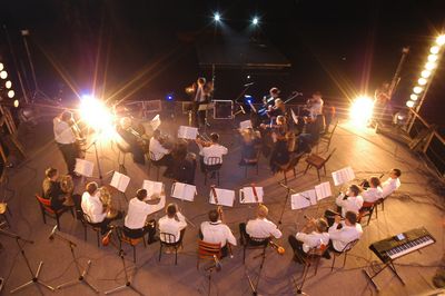 Abbildung weiß gekleidete Musiker sitzen im Halbkreis in einem beleuchtetem Orchestergraben