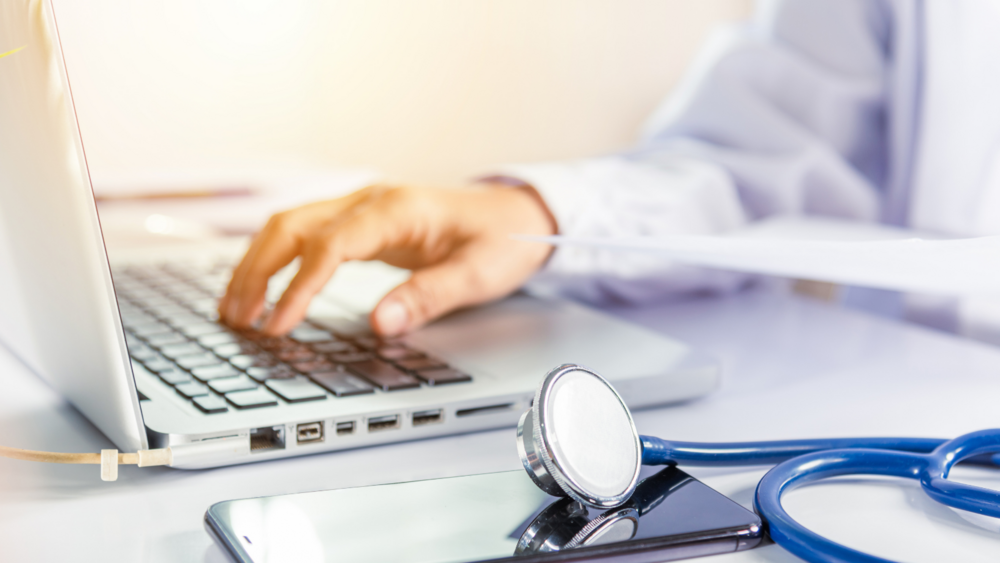 Abbildung ein Arzt sitzt an seinem Schreibtisch mit einem Laptop und arbeitet daran, daneben liegt ein Stethoskop