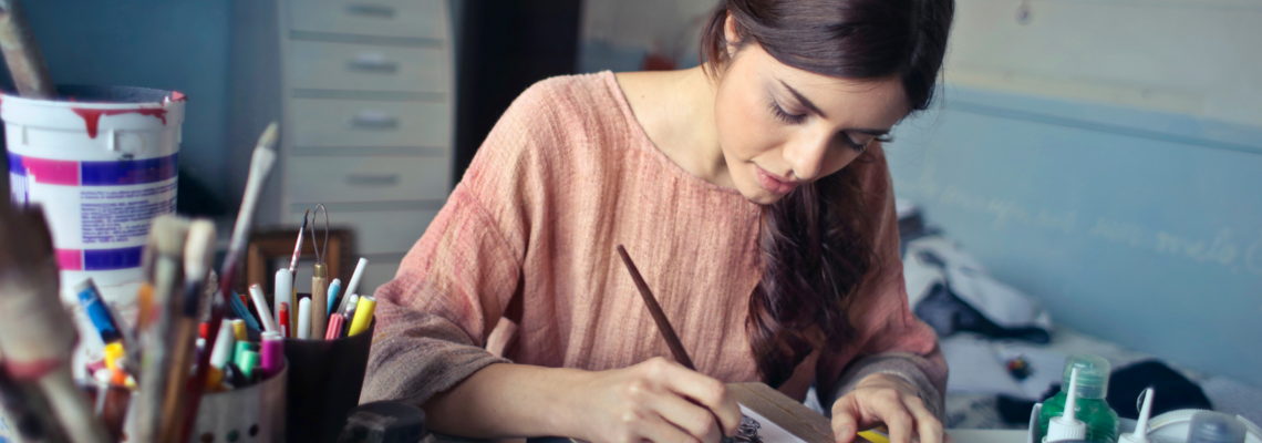 Abbildung eine jugendliche Frau sitzt an einem Schreibtisch und zeichnet mit einem Pinsel ein Bild