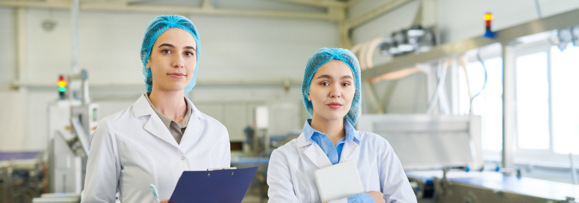 Abbildung zwei Frauen in hygienischer Schutzkleidung stehen in einem Labor und schauen in die Kamera