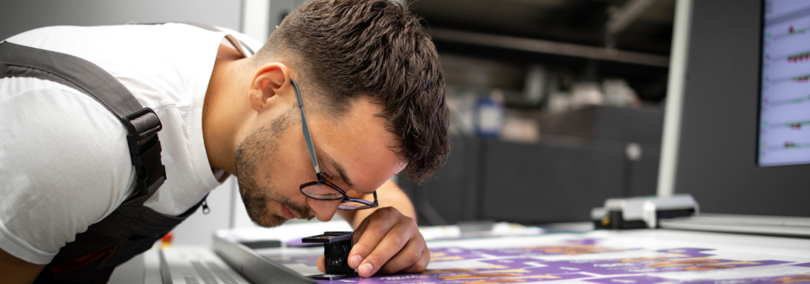 Abbildung Ein Mann schaut mit einer Lupe auf einen Frischen druck in einen Industriedrucker 