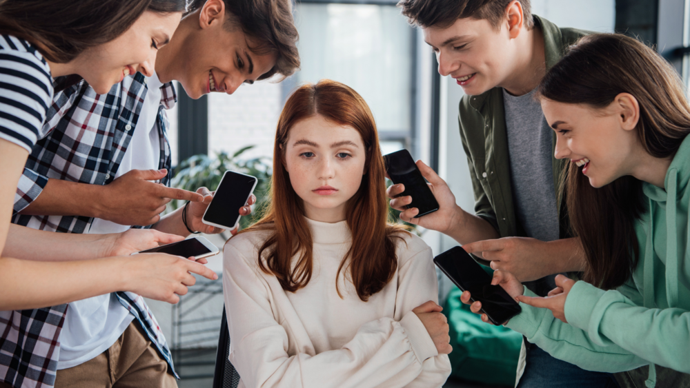 Abbildung ein rothaariges Mädchen wird von vier Jugendlichen umkreist, sie schaut traurig, während die anderen vier lachen und auf deren Handys zeigen.