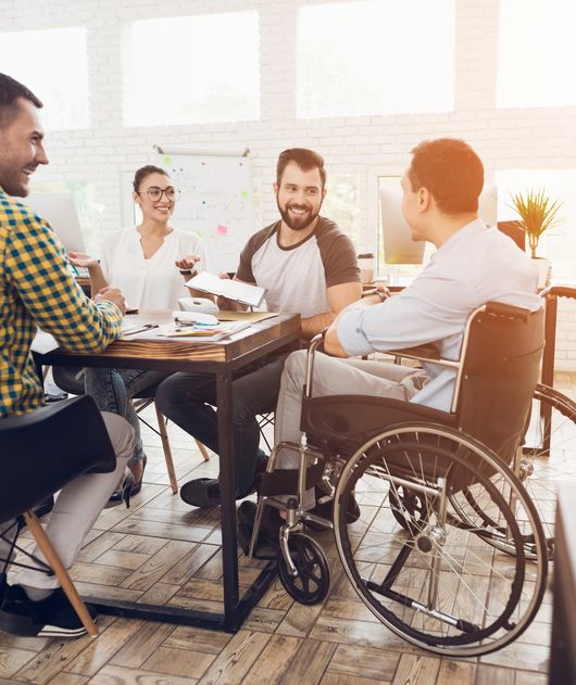 Abbildung Person im Rollstuhl sitzt an einem Tisch mit anderen Personen