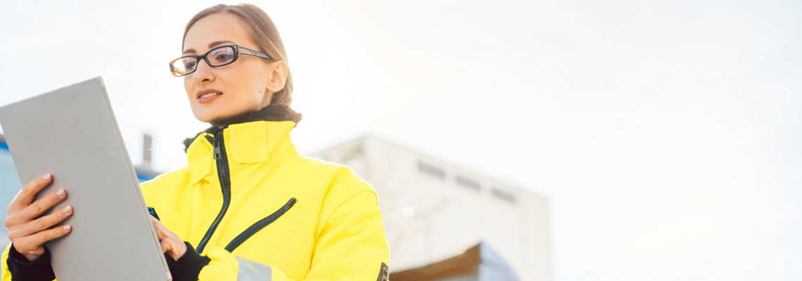 Abbildung Frau steht mit gelber Schutzjacke auf der Baustelle mit Tablet 