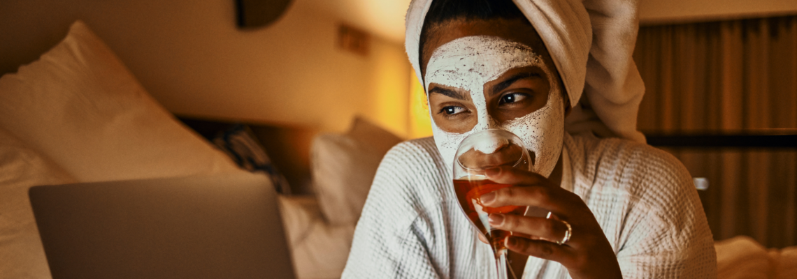 Abbildung eine Frau liegt mit Gesichtsmaske, Handtuch in den Haaren und Bademantel auf einem Bett während sie etwas aus einem Weinglas trinkt und in einen Laptop schaut 