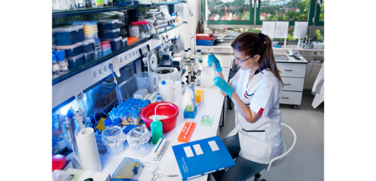 Abbildung junge Frau mit weißem Kittel und blauen Handschuhen sitzt am Labortisch