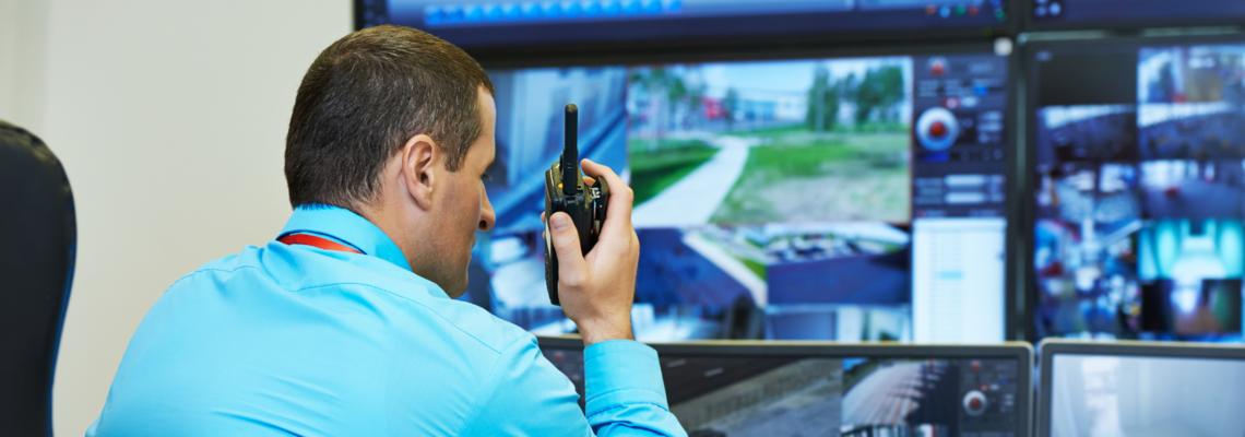Abbildung ein Mann schaut auf Bildschirme von Überwachungskamerads und spricht in ein Funkgerät 