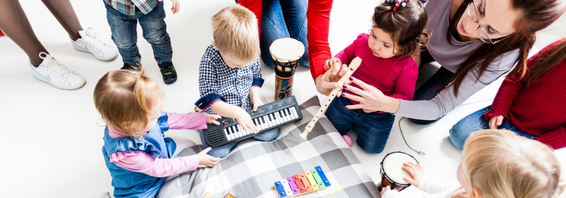 Abbildung Kleinkinder spielen mit Instrumenten auf einem Kissen drumherum stehen die Eltern 