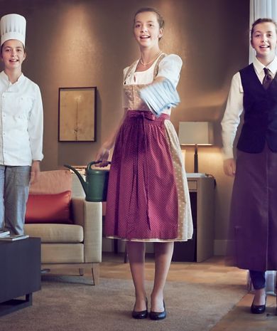 Abbildung Frau im Dirndl mit Gießkanne zwischen Köchin und Kellnerin stehend in einem Hotelzimmer