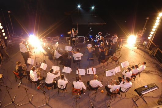 Abbildung weiß gekleidete Musiker sitzen im Halbkreis in einem beleuchtetem Orchestergraben