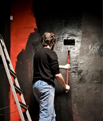 Abbildung Mann mit malt mit Malerrolle an einer schwarz-roten Wand. Daneben eine Leiter