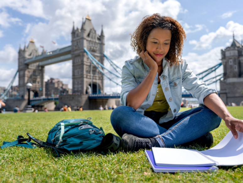 Abbildung eine junge Frau sitz auf einer Wiese vor der London Bridge und lernt