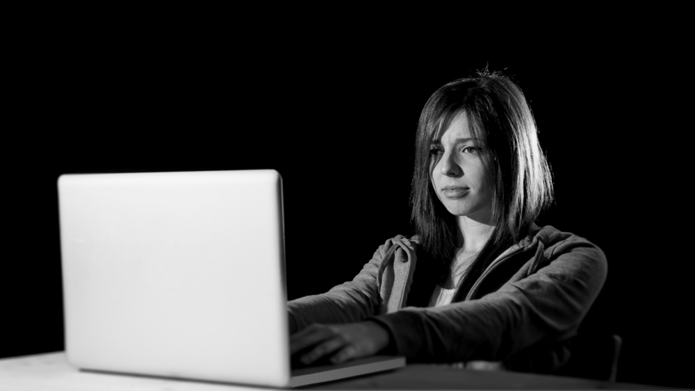 Abbildung in schwarz und weiß von einem Mädchen, dass mit verzogenen Gesicht hinter ihrem Laptop sitzt.