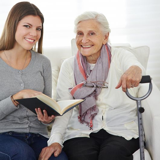 Abbildung Junge Frau mit Buch in der Hand sitzt neben einer alten Frau, die eine Gehhilfe in der Hand hält