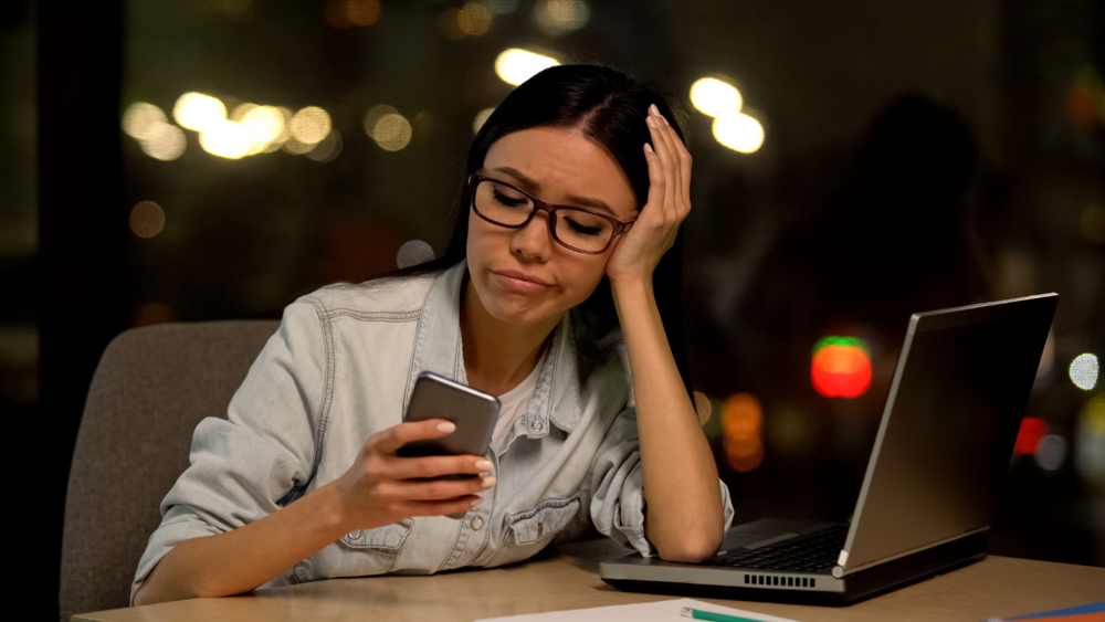 Abbildung eine Jugendliche sitzt an ihrem Schreibtisch und hat ihr Handy in der Hand, während sie sich auf ihren aufgelehnten Laptop abstützt.