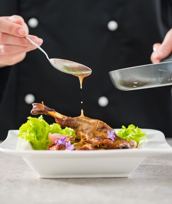 Abbildung Person gibt aus einem Topf mit einem Löffel Sauce auf ein Gericht